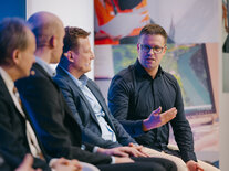 Ein Teilnehmender der Diskussion auf der Bühne erläutert einen wichtigen Punkt, drei andere Teilnehmende sitzen ihm zugewandt da