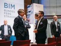 Zwei Männer im Gespräch, im Hintergrund weitere Teilnehmer des Fachsymposiums vor Schriftzug "BIM BUNDESFERNSTRASSEN"