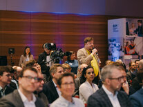 Ein Teilnehmender im Publikum mit braunem Jacket und gelbem Hemd spricht seine Frage in das Mikrofon im Saal