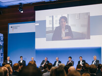 Auf dem Großbildschirm über dem Podium wird Vera Grote, die Moderatorin des Online-Teils des Fachsymposiums eingeblendet, die Fragen aus dem Chat weitergibt