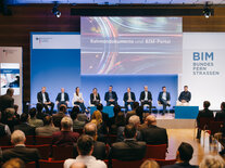Podiumsdiskutanten auf der Bühne, auf dem Großbildschirm läuft der Trailer für die Session "Rahmendokumente und BIM-Portal"