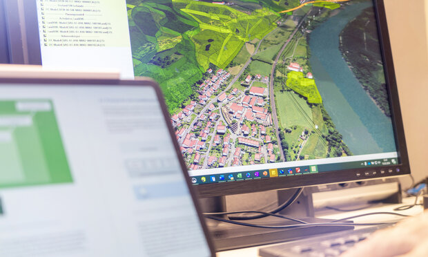 Auf einem Bildschirm ist eine Luftaufnahme eines Wohngebietes zu sehen, das von Feldern und einem Fluss umgeben ist. Im Vordergrund ist unscharf ein Tablet zu erkennen. 