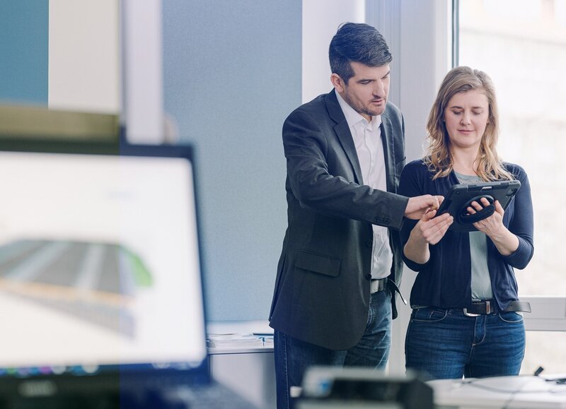 Zwei Mitarbeitende in einem Büro betrachten gemeinsam ein Tablet, während auf einem Bildschirm im Vordergrund ein digitales Modell zu sehen ist.