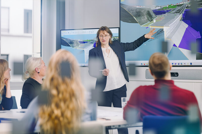 Eine Frau steht vor einem großen Monitor, auf dem ein BIM-Modell zu sehen ist. Sie erläutert etwas am Modell, die Zuschauer im Vordergrund hören aufmerksam zu.