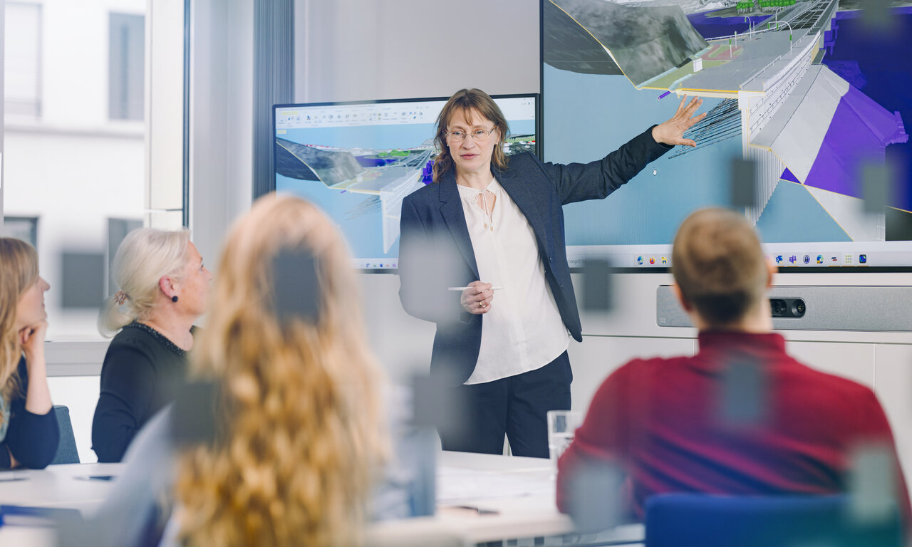 Eine Frau steht vor einem großen Monitor, auf dem ein BIM-Modell zu sehen ist. Sie erläutert etwas am Modell, die Zuschauer im Vordergrund hören aufmerksam zu.