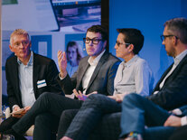 Drei Teilnehmer des Podiums "Pilotprogramm und Reifegradmessung" diskutieren, der Moderator hört zu