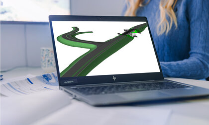 Ein Laptop steht auf einem Schreibtisch. Der Bildschirm zeigt eine BIM-generierte Darstellung einer Straße mit Unterführung. Im Hintergrund sind ein Büro, ein größerer Monitor an der Wand und eine Mitarbeiterin im blauen Pullover zu erkennen.