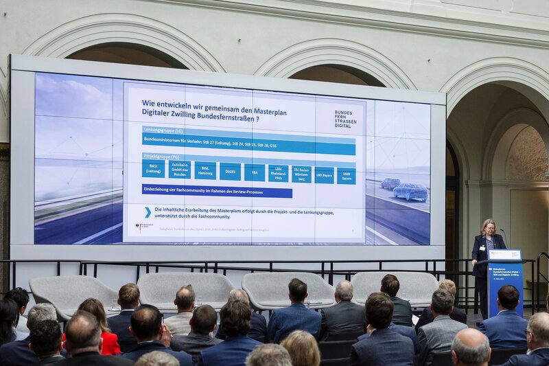 Vortrag beim Dialogforum Bundesfernstraßen Digital: Eine Referentin steht am Rednerpult vor einem großen Publikum. Auf der Leinwand ist eine Übersicht zur gemeinsamen Entwicklung des Masterplans Digitaler Zwilling Bundesfernstraßen zu sehen.
