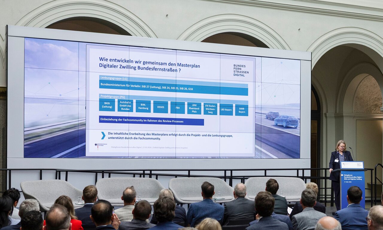 Vortrag beim Dialogforum Bundesfernstraßen Digital: Eine Referentin steht am Rednerpult vor einem großen Publikum. Auf der Leinwand ist eine Übersicht zur gemeinsamen Entwicklung des Masterplans Digitaler Zwilling Bundesfernstraßen zu sehen.