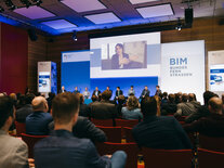 Publikum von hinten vor der Bühne, auf dem Großbildschirm gibt die Chat-Moderatin die Fragen der Online-Teilnehmenden in den Saal