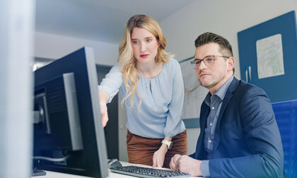 Zwei Personen im Büro schauen gemeinsam auf einen Computerbildschirm; die Frau zeigt auf den Monitor, der Mann sitzt daneben und hört aufmerksam zu. 