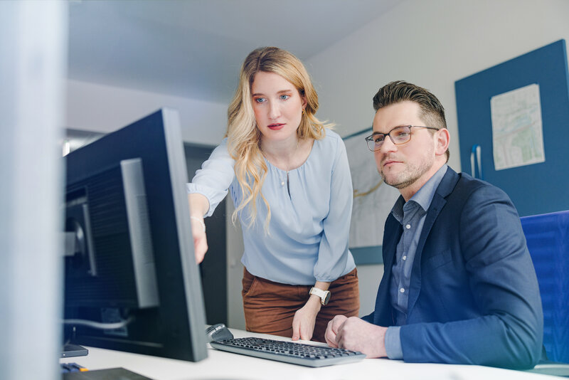 Zwei Personen im Büro schauen gemeinsam auf einen Computerbildschirm; die Frau zeigt auf den Monitor, der Mann sitzt daneben und hört aufmerksam zu. 