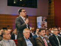 Ein Teilnehmer im grauen Anzug diskutiert aus dem Publikum heraus mit dem Podium