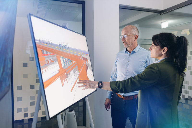 Ein Mann und ein Frau stehen vor einem Großbilschirm, die Frau deutet auf die BIM-Visualisierung eines Brückenbauwerks