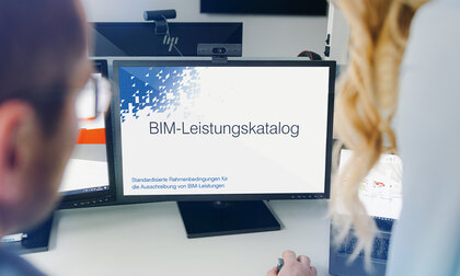 Zwei Personen blicken auf einen Monitor, auf dem das Titelblatt des BIM-Leistungskatalogs gezeigt wird.